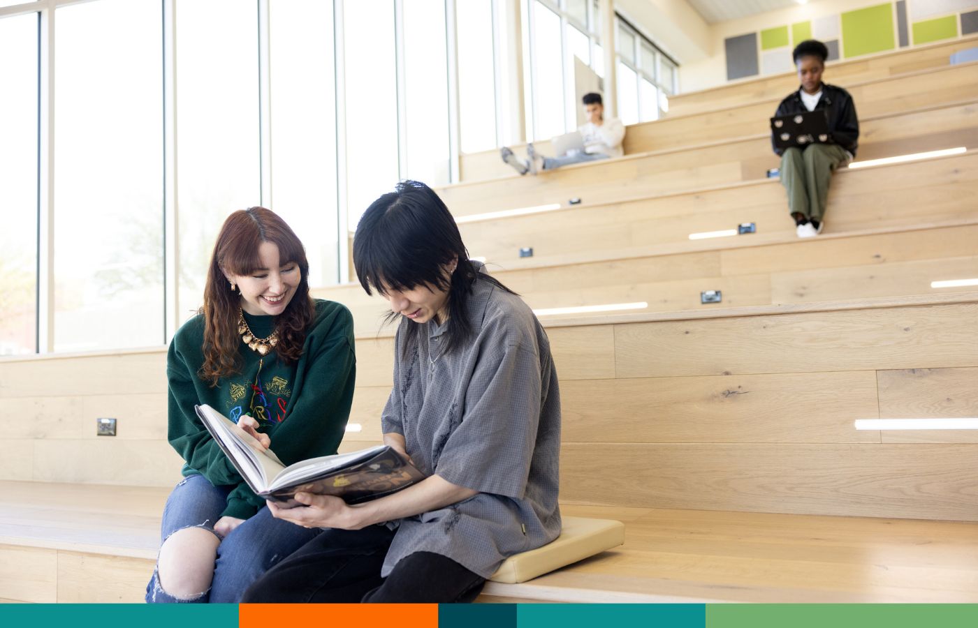 Two students sitting in a common space looking at a book together and laughing