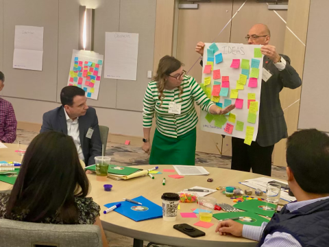 People at a conference table. I woman is pointing at post-it notes on a large paper that is being held up by another person
