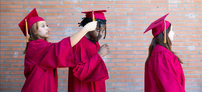 Three students with graduation regalia on. one girl adjusts the string on top of her classmates hat.