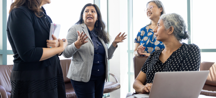 Four women in a meeting room engage in discussion around a table. One woman gestures while speaking as others listen, with one seated participant working on a laptop near large windows.