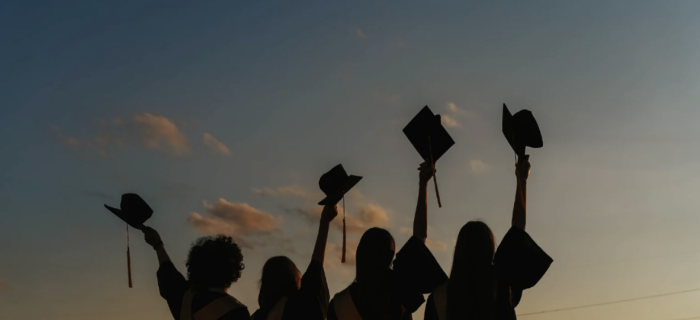Four silhouettes of students holding their graduation caps up during sunset.